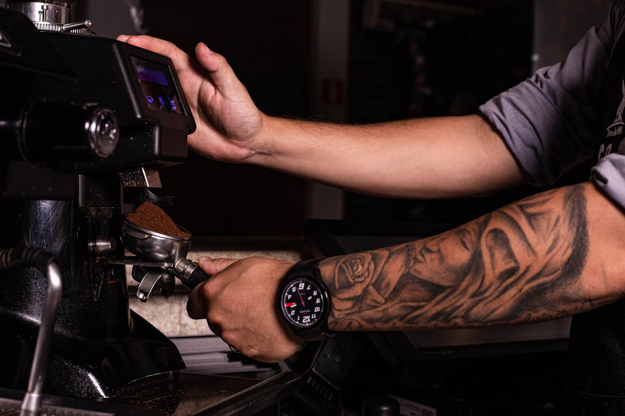 hero-img Close-up of a baristas tattooed arm using an espresso machine to grind coffee.